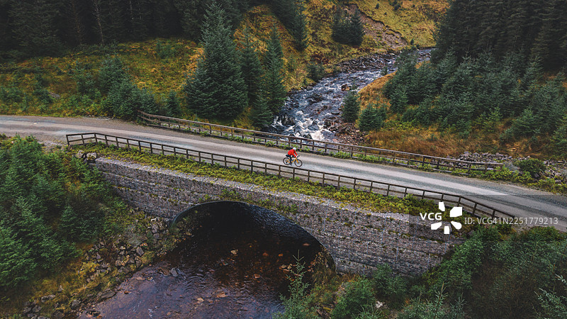 苏格兰偏远森林中，无人机/空中视角拍摄的成熟男子在河上砂石小径骑行。图片素材