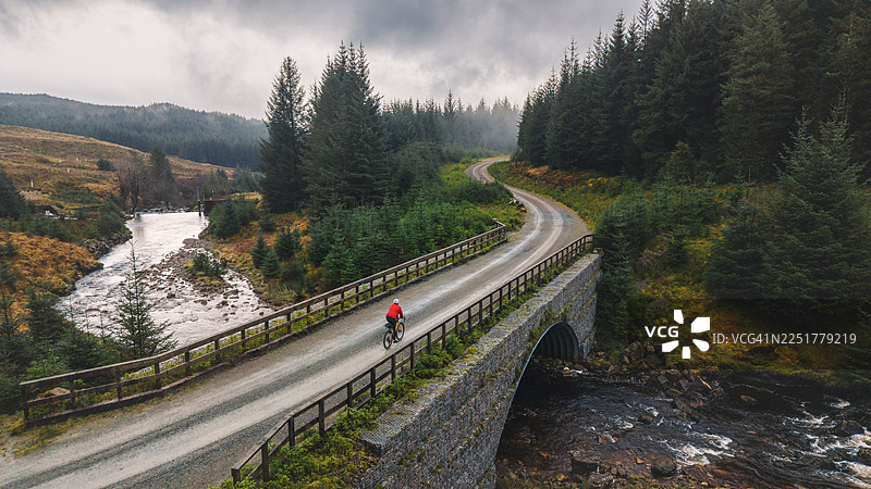 苏格兰偏远森林中，无人机/航拍视角下，一位成熟男子在河上砾石小径上骑行图片素材