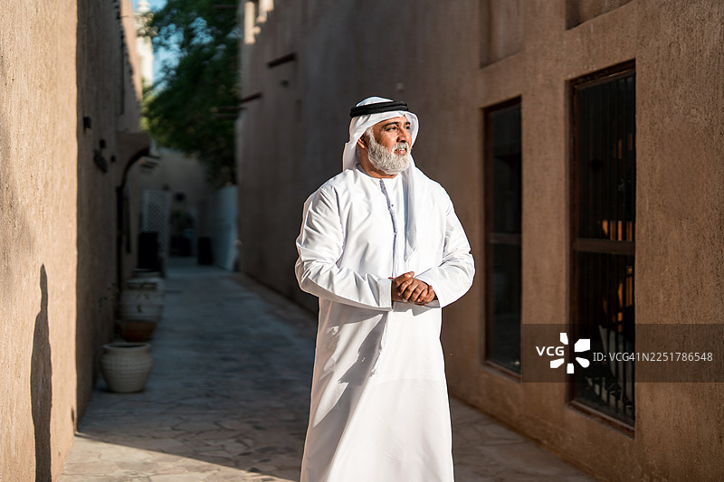 一位穿着白色坎杜拉、神情庄重的年长阿联酋男子，在传统的中东小巷中微笑。图片素材