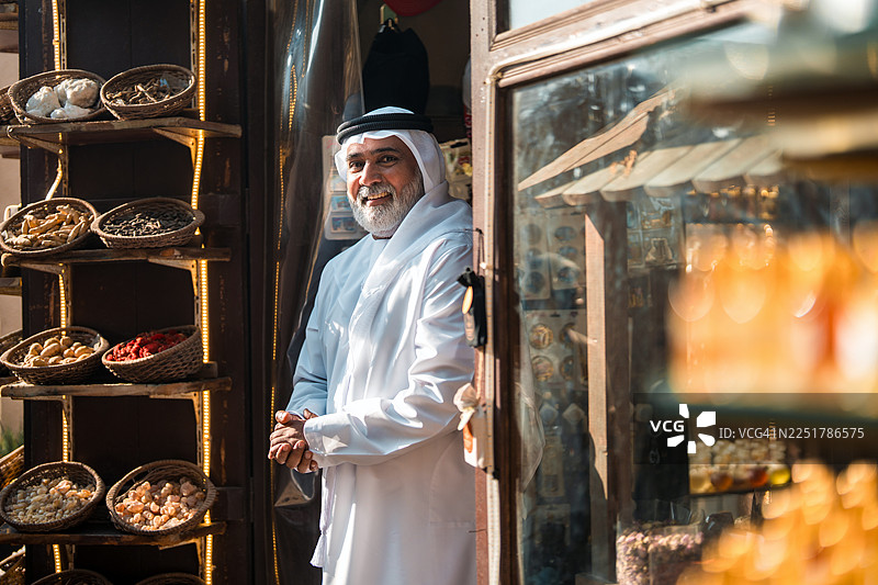 一位身穿传统白色坎杜拉的快乐阿拉伯老年商人，站在历史悠久的店铺门口图片素材