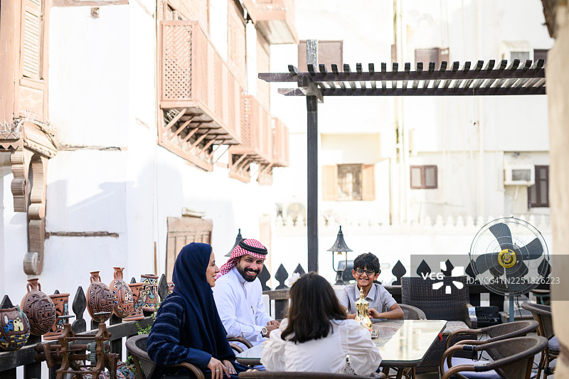 阳光明媚的庭院里，一家人坐在户外的咖啡馆桌旁，背景是传统建筑，广角镜头拍摄，他们看起来很高兴。图片素材