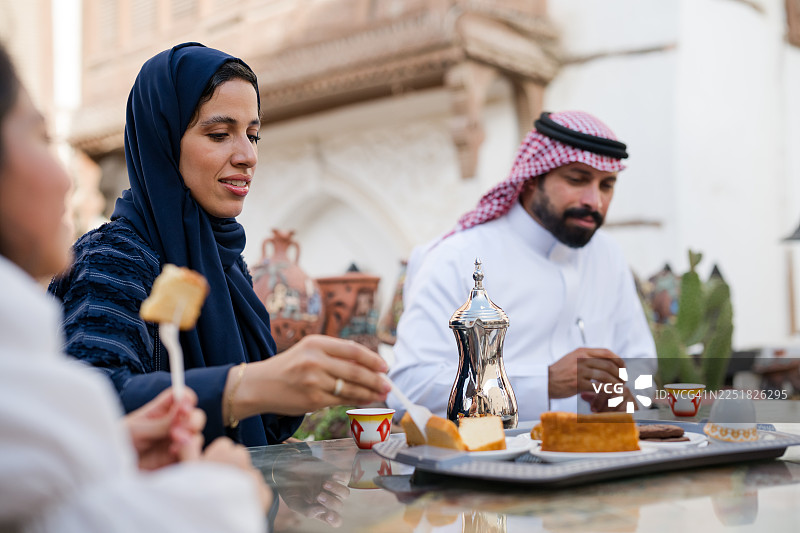 一位戴着头巾、身穿阿巴亚的母亲在户外咖啡馆的餐桌旁与家人一同坐着，手中拿着甜点，背景是历史街区。图片素材