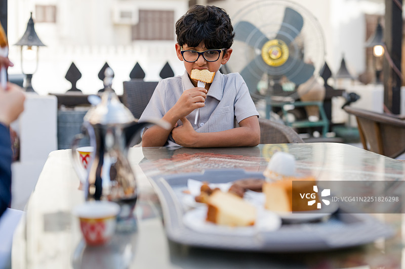 户外咖啡馆里，一个年轻的阿拉伯男孩在桌边吃着蛋糕，前景是糕点，抓拍瞬间。图片素材