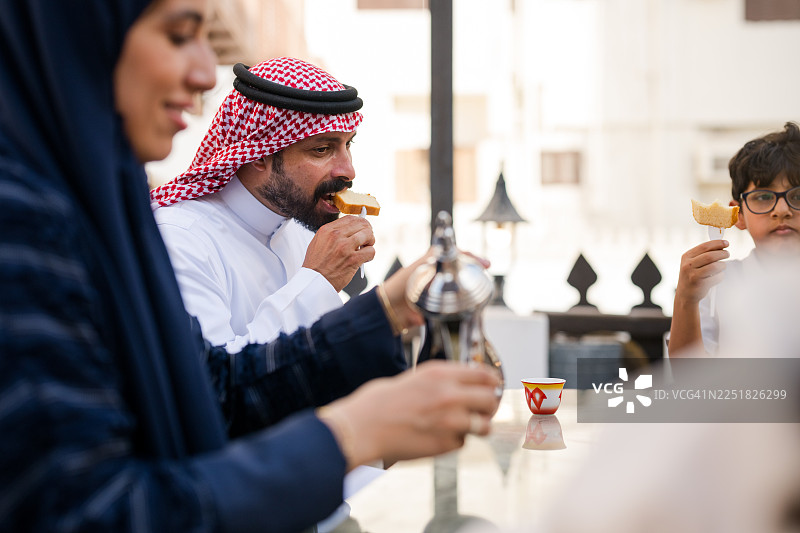 阿拉伯母亲和父亲身着传统服饰，在户外咖啡馆的餐桌旁与家人一同享用甜点，背景是历史街区，侧面视角。图片素材