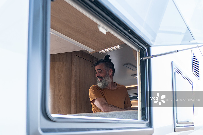 留着胡子的男人微笑着，望着露营车窗外，享受着旅行冒险中的自由与开阔的道路图片素材