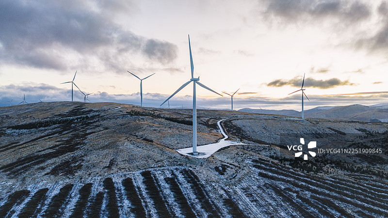 在寒冷的冬日日落时分，苏格兰高地山坡上风力涡轮机的宽广航拍/无人机视角图片素材