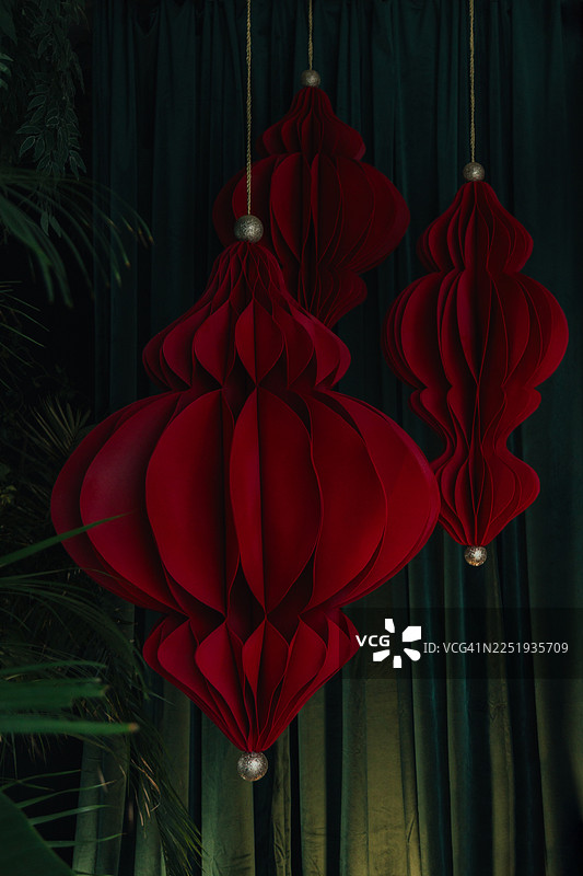 红色的纸蜂巢装饰品用细绳挂着，靠近绿植，背景是深绿色窗帘。图片素材