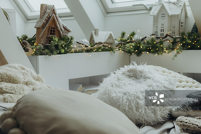 一个节日气氛的阁楼角落，铺着柔软的床铺，松软的房屋和闪烁的灯光点缀在常绿树枝中。图片素材