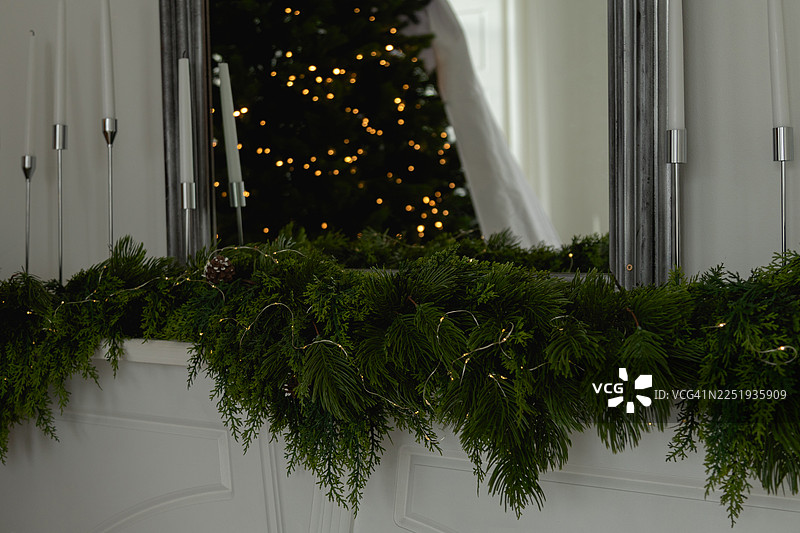 一个白色的壁炉架，装饰着常青藤花环、串灯、高蜡烛的锥形烛台，以及一面镜子，镜中映照出圣诞树的璀璨灯光。图片素材