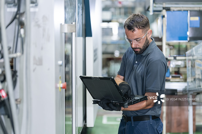 技术人员在工厂使用加固型计算机操作塑料注塑机控制面板，以验证设置的正确性。图片素材