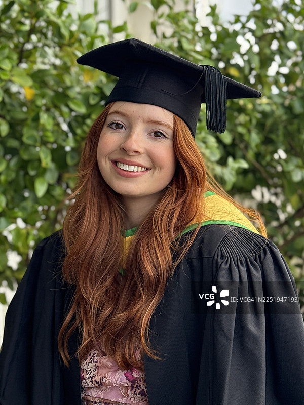 一位迷人的红发女研究生身穿黑色短袖毕业礼袍，内搭粉色碎花连衣裙，佩戴绿色和黄色帽兜，头戴缀有流苏的黑色学士帽，进行正面肖像拍摄，焦点在前景。图片素材