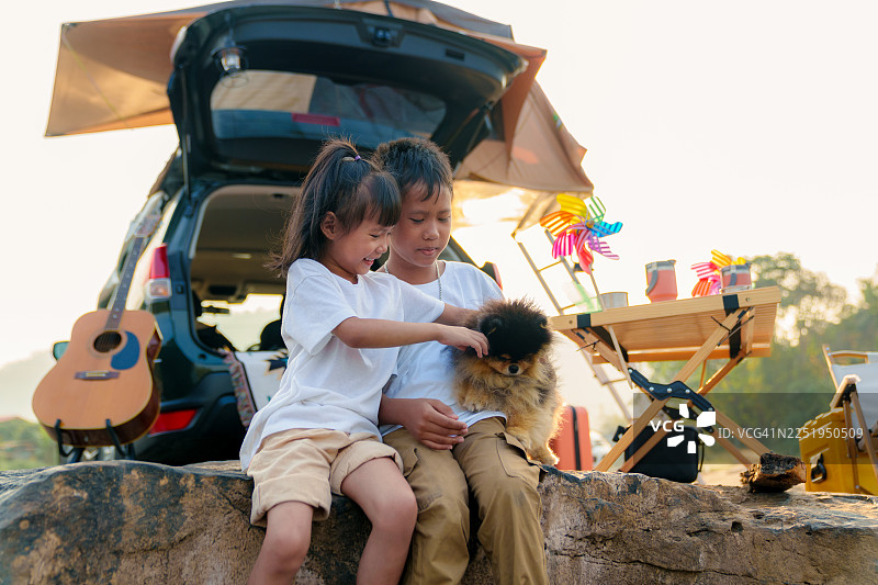 两个开心的孩子和他们的宠物博美犬，在家庭户外度假期间，坐在一辆配有车顶帐篷的车旁边的岩石上。图片素材