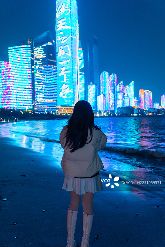 一位年轻女子在中国山东青岛市，在青岛第三海水浴场上空眺望灯火辉煌的青岛城市天际线。图片素材