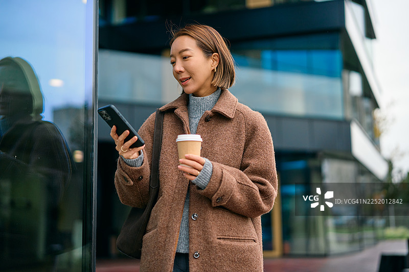 一位女士在城市里一边走一边喝咖啡，同时使用手机。图片素材