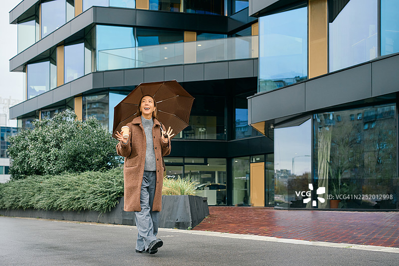 年轻的女子带着咖啡和雨伞走在城市里，看起来很高兴图片素材