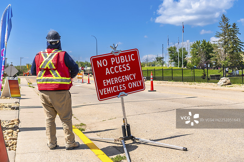 晴天在阿尔伯塔省德拉姆赫勒，一名建筑工人戴着安全背心和安全帽，站在禁止公众进入的标志附近，为紧急车辆控制道路通行。图片素材
