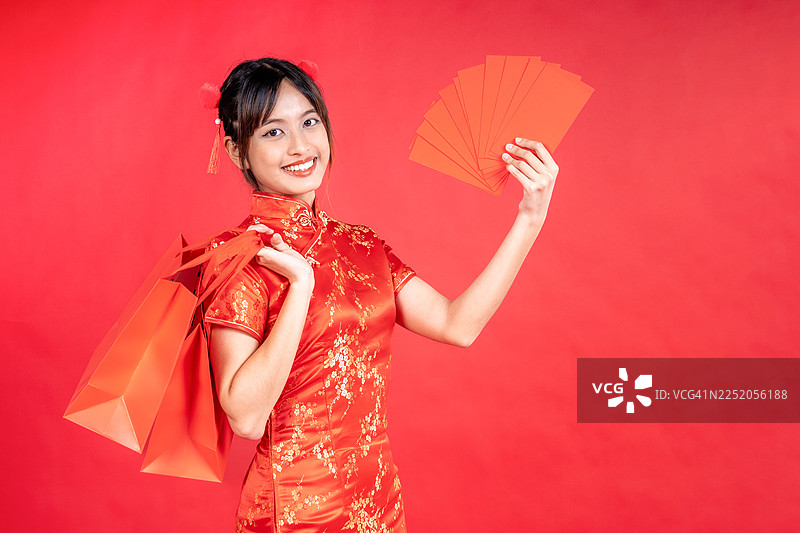 穿着传统中式旗袍的快乐亚洲购物狂女人，手持购物袋和红包，背景为红色，新年快乐。图片素材