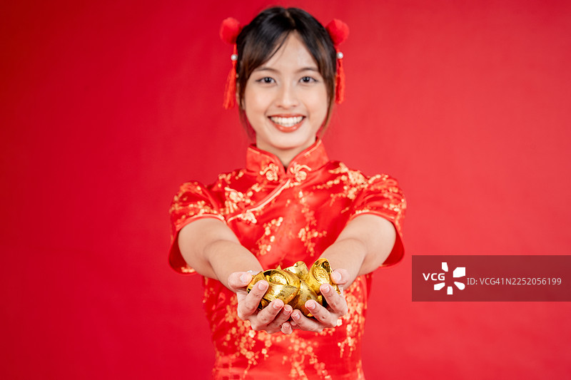 新年快乐。一位身着传统旗袍的亚洲女性，手持象征财富和繁荣的金元宝，背景为红色。图片素材