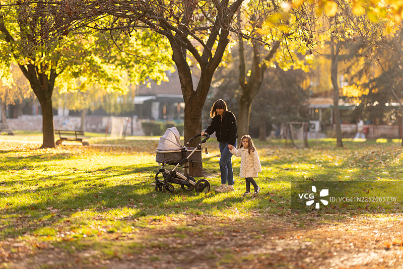 阳光明媚的秋日公园里，一位母亲推着婴儿车，两个孩子在旁边走着图片素材