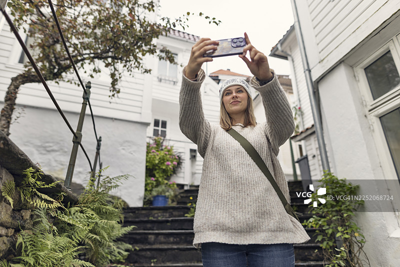 年轻女子漫步在卑尔根宁静的小巷迷宫中，周围是白色外墙和盆栽绿植。图片素材