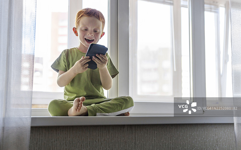 开心的男孩在家坐在窗台上玩手机图片素材