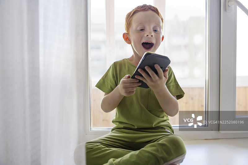 红发男孩在家坐在窗边，享受着使用智能手机的时光图片素材