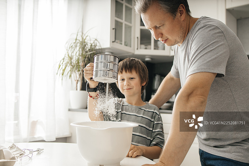 父亲站在厨房的桌子旁，儿子在碗里筛面粉图片素材