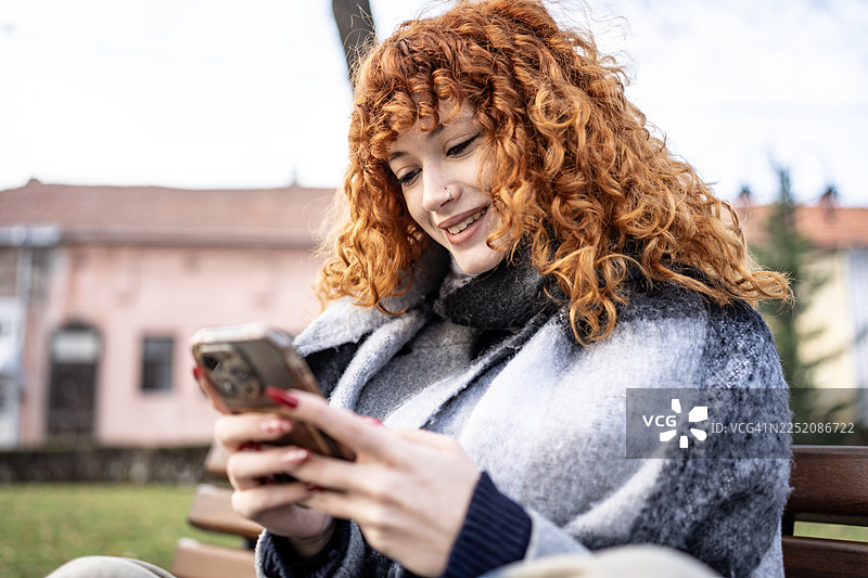 红发女子坐在公园长椅上，面带微笑地看着手机，置身于舒适的户外环境中图片素材