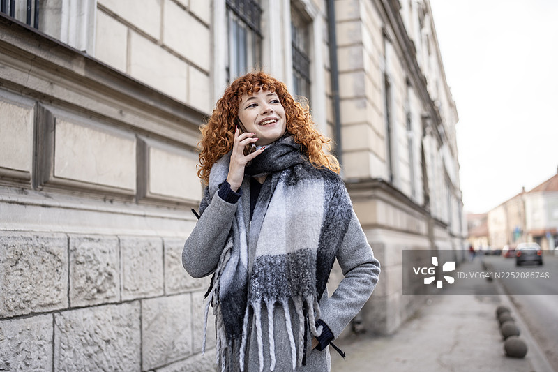 身穿灰色外套和围巾的红发女子在城市街道上微笑着打电话图片素材
