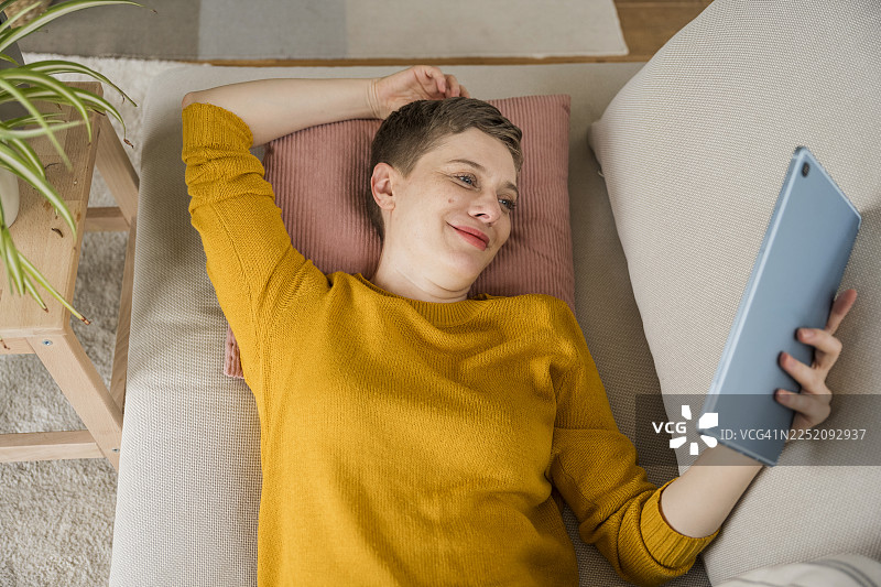 女人在家放松地躺在沙发上，面带微笑地使用平板电脑图片素材