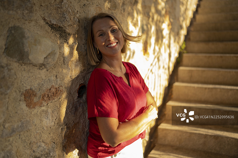 阳光下，一名女子面带微笑，倚靠在室外楼梯旁的墙壁上。图片素材