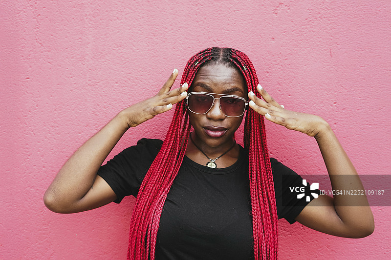 年轻女子留着红色辫子，戴着太阳镜，在粉色墙壁前摆姿势图片素材