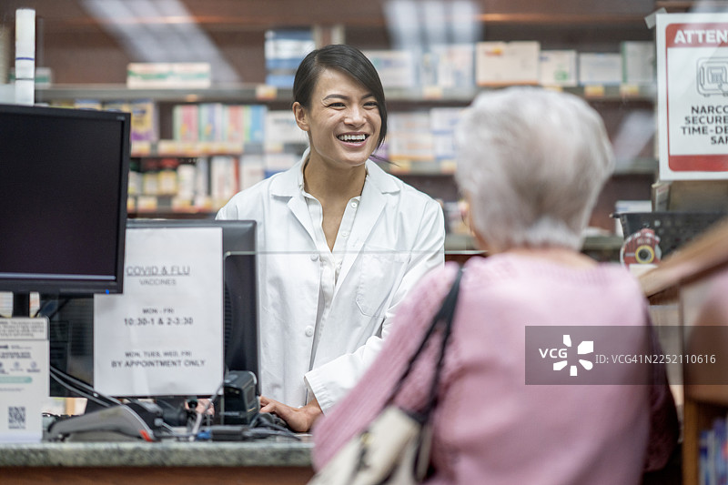 一位微笑的亚洲女药剂师在繁忙的药房柜台协助一位老年顾客图片素材