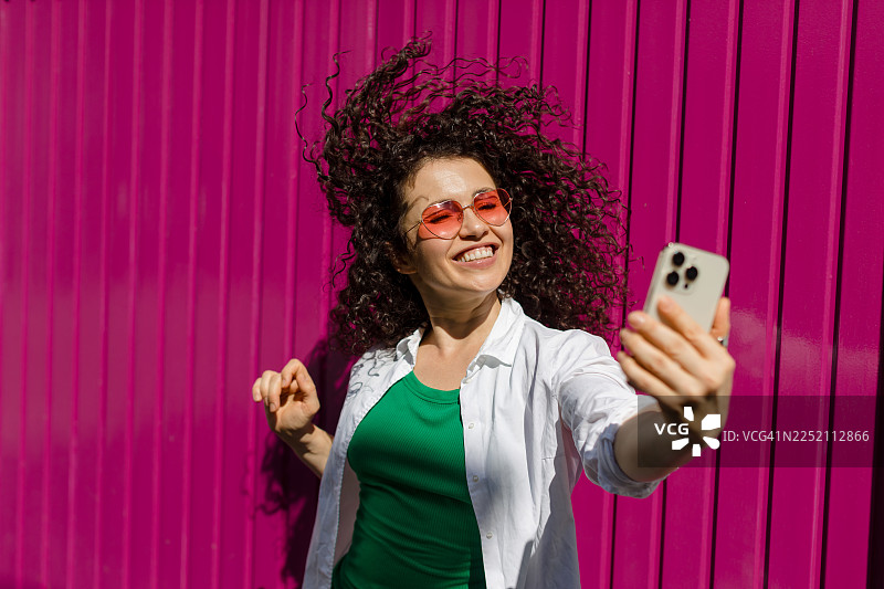 一位成年人正拿着智能手机在粉色墙前自拍，脸上带着笑容图片素材