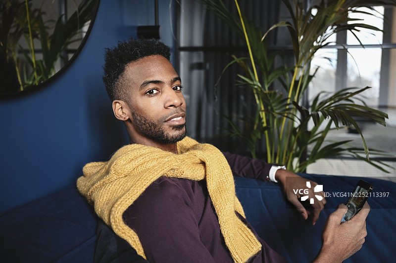 穿着黄色毛衣的男人坐在室内，使用智能手机看向镜头图片素材