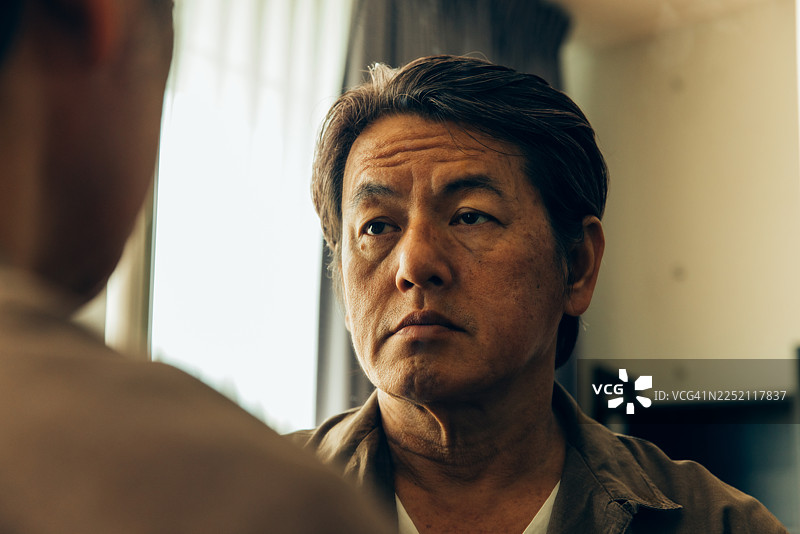 下午，一个男人坐在房间里，窗户旁边，柔和的光线下，看着自己的倒影。图片素材