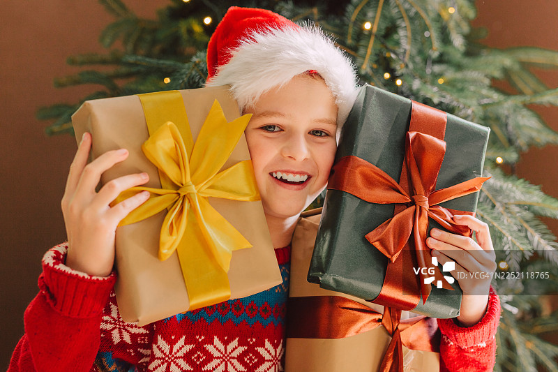 戴着圣诞帽的微笑儿童在室内树旁拿着圣诞礼物图片素材