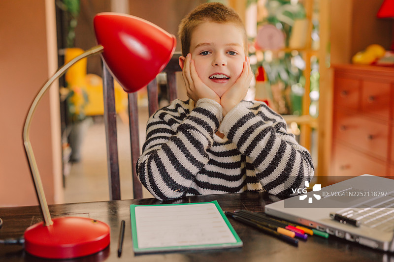 一个男孩坐在室内桌子旁，用笔记本电脑做作业，看起来很疲倦图片素材