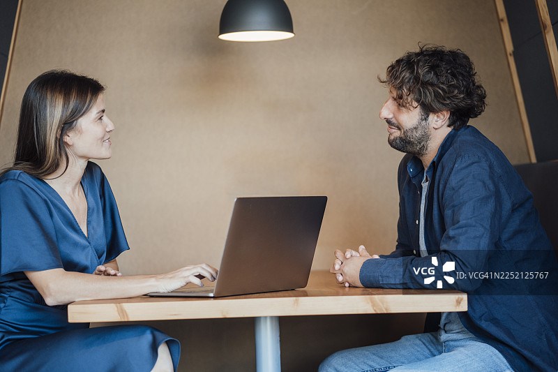 快乐的女商人坐在办公室隔间里，一边使用笔记本电脑一边与男商人交谈图片素材