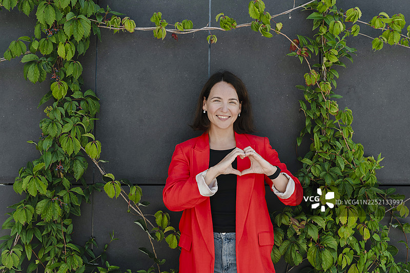 身穿红色西装的女商人在户外植物背景下做出心形手势图片素材