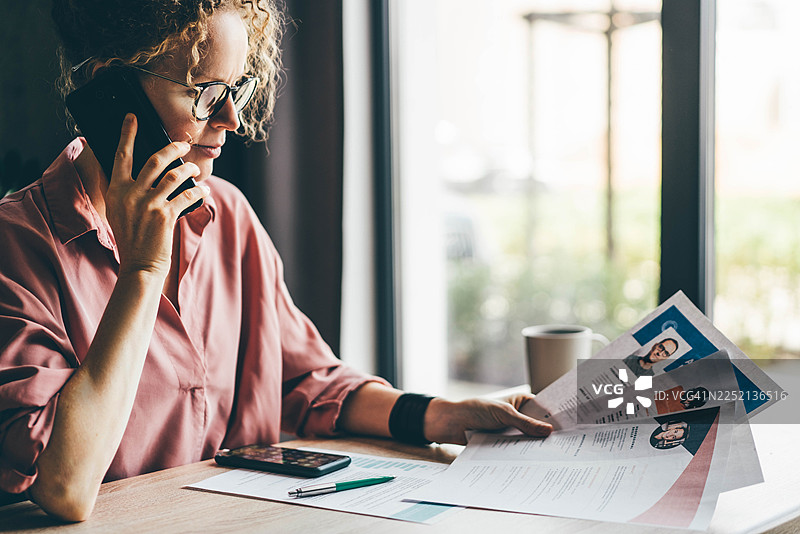 一位戴眼镜的专注女性在明亮的办公室里，一边对着智能手机通话，一边审阅打印的简历。这一场景代表了招聘、人力资源和现代专业的招聘流程。图片素材