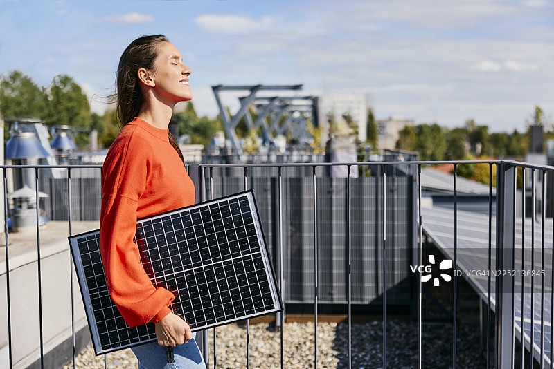 一位快乐的女士站在屋顶露台上，手里拿着一块太阳能电池板图片素材