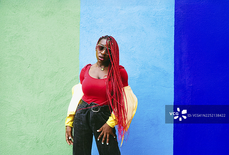 一位时尚女子留着红色编发，在户外的彩色墙壁旁摆姿势拍照。图片素材