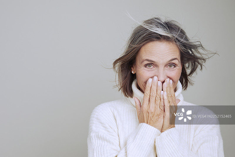 一位穿着舒适高领衫的女士，用手捂着嘴，脸上带着微笑，这是一幅肖像照图片素材