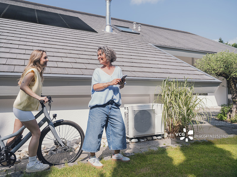 母女俩骑着自行车，在一栋带有太阳能电池板和热泵的房子附近交谈图片素材
