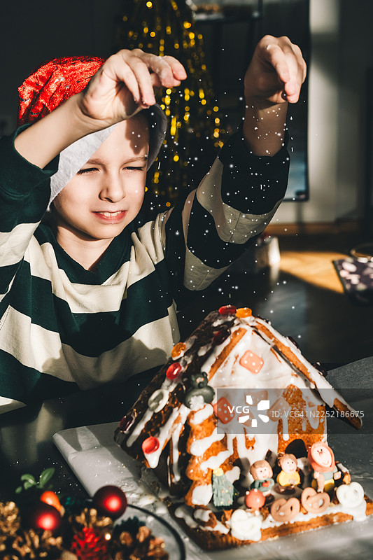 一个戴着圣诞帽的孩子在节日气氛浓厚的圣诞桌旁，用糖霜装饰自制的姜饼屋，在灯光、装饰和温馨的家庭传统中营造出温暖的节日氛围。图片素材