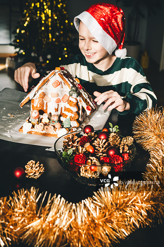 戴着圣诞帽的孩子在节日圣诞桌旁用糖霜装饰自制的姜饼屋，营造出灯光、装饰和温馨家庭传统营造的温暖节日气氛。图片素材