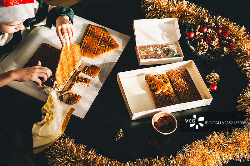 戴着圣诞帽的孩子在节日餐桌旁用糖霜装饰自制的姜饼屋，营造出灯光、装饰和温馨家庭传统交织的温暖节日氛围。图片素材