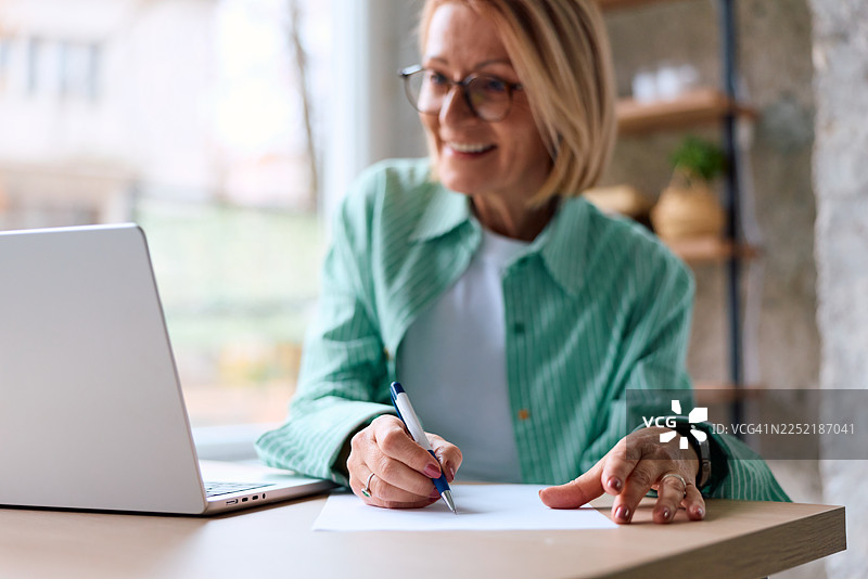 一位成熟的成年女性正在笔记本上写笔记，同时对着笔记本电脑微笑图片素材