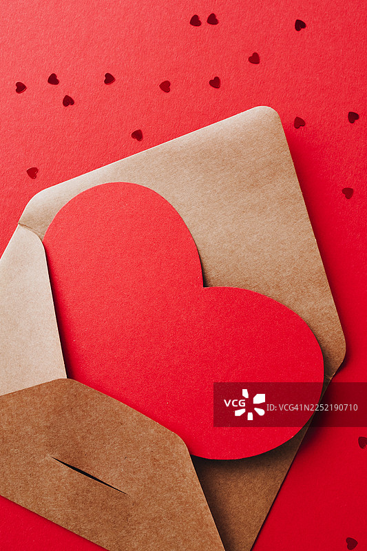 竖直特写俯视，一个红色的纸质心形物品，放在一个打开的牛皮纸信封里，背景是鲜亮的红色，上面散落着细小的爱心纸屑。图片素材